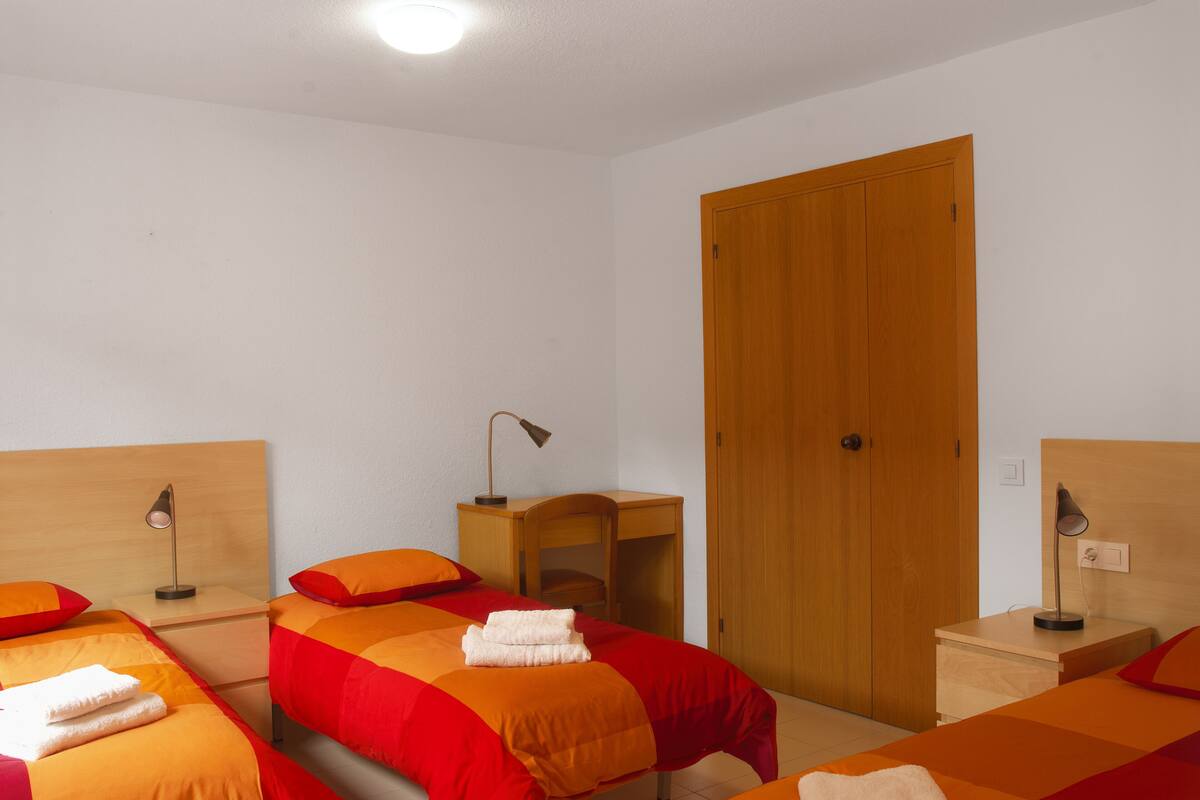 A hotel room with two beds, wooden headboards, a wooden wardrobe, a nightstand with a lamp, and a small wooden table.