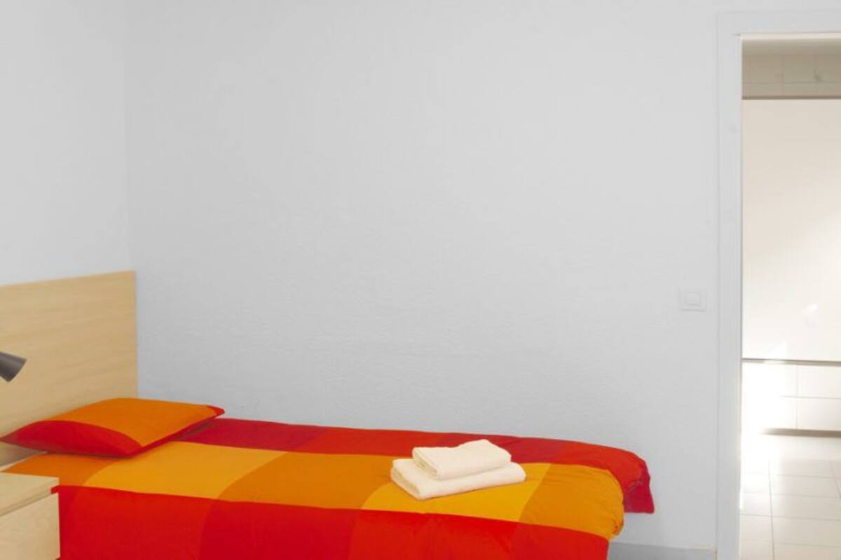 A single bed with a red and yellow striped bedspread, a white pillow, and a folded white towel on the bed. There is a bedside table with a lamp and a power outlet on the wall.