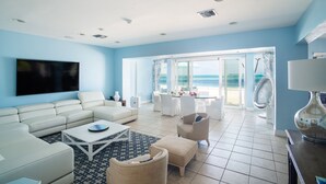 A spacious living room with a large flat-screen TV, white furniture, and a view of the ocean through large glass doors. - LUXURY HOUSE WITH PRIVATE BEACH (Nassau)