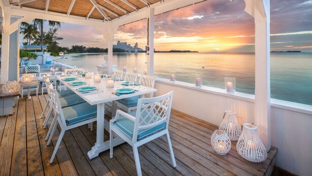 A covered outdoor dining area with white furniture and teal cushions, overlooking a body of water and a sunset. - LUXURY HOUSE WITH PRIVATE BEACH (Nassau)