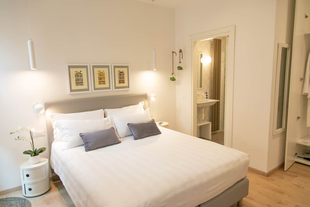 A neatly arranged bedroom with a bed, bedside tables, a nightstand, a potted plant, framed pictures on the wall, and a bathroom visible through an open door.
