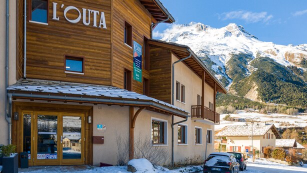 Un bâtiment en bois avec une enseigne indiquant « L’OUTA », et en arrière-plan, une vue sur des montagnes enneigées.