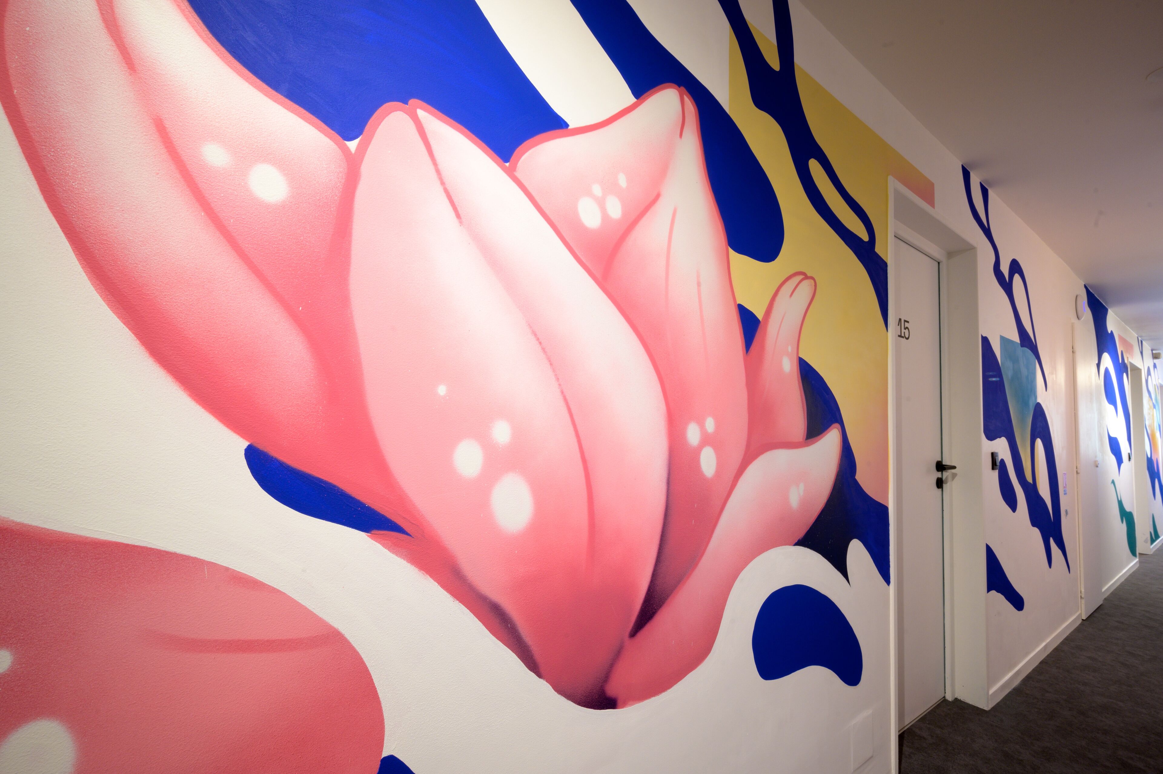 Un pasillo con un mural floral llamativo en la pared, una puerta con el número 15, y suelo alfombrado.