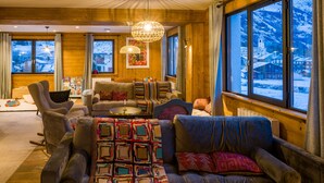 Un intérieur chaleureux avec des murs en bois, une grande fenêtre donnant sur un paysage enneigé et un espace salon central agrémenté d’un plaid à motifs.