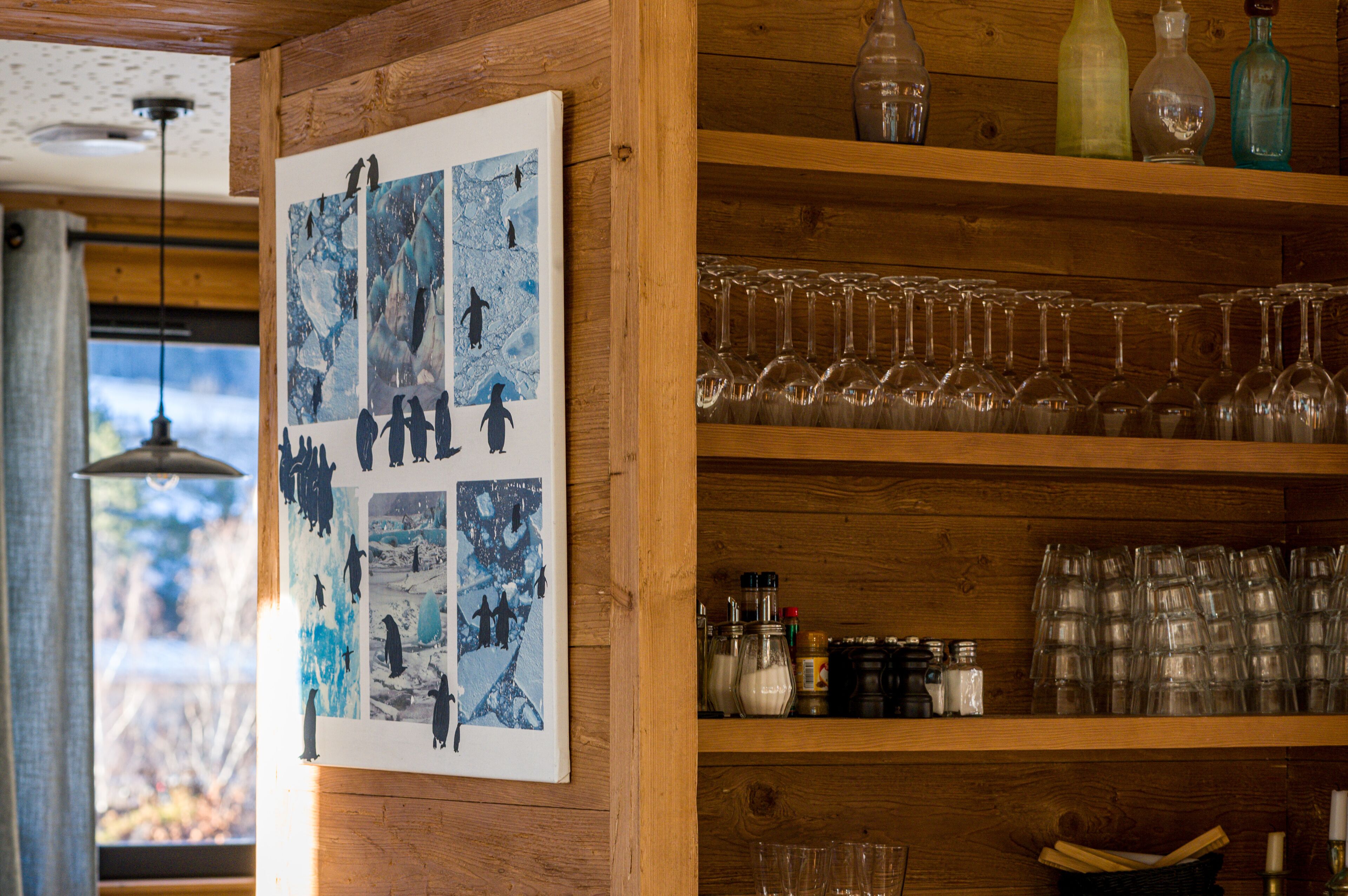 Un intérieur en bois, avec une affiche de pingouins accrochée au mur, des étagères remplies de verres à vin et une fenêtre donnant sur des arbres.