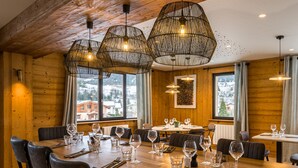Un intérieur en bois avec une table à manger dressée pour six personnes, entourée de chaises, et de grandes fenêtres avec des rideaux.