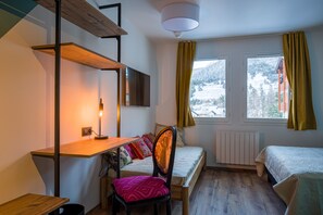 Une petite pièce comprenant un lit, un bureau et une chaise. On y trouve une fenêtre avec des rideaux, un radiateur et une vue sur un paysage enneigé.