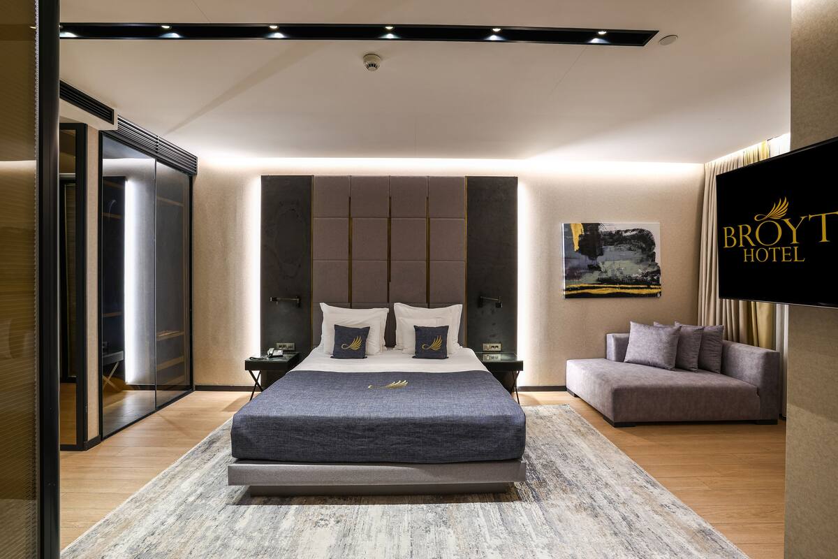 A modern hotel room with a large bed, a sofa, a flat-screen TV, and a wall-mounted artwork.