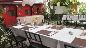 Un área de comedor al aire libre con mesas dispuestas para dos, cubierta por un toldo blanco, rodeada de follaje y un edificio rojo al fondo.