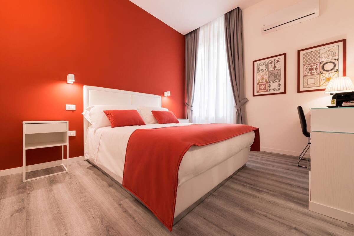 Une chambre d’hôtel moderne avec un mur d’accent rouge, un lit blanc avec des oreillers rouges, une petite table de chevet blanche, un bureau avec une chaise et deux tableaux encadrés accrochés au mur.