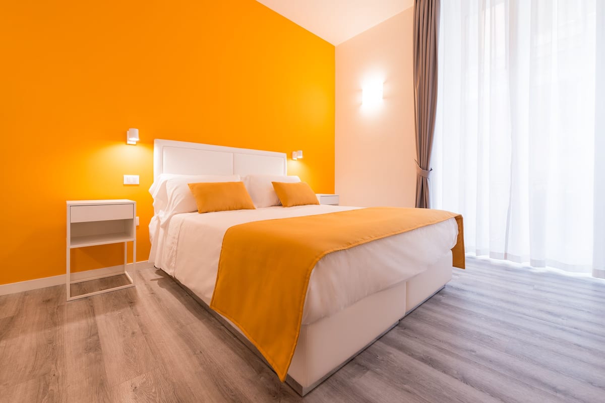 Une chambre d’hôtel moderne avec un mur orange vif, un lit blanc agrémenté d’un plaid jaune, un plancher en bois et une table de chevet avec une lampe.