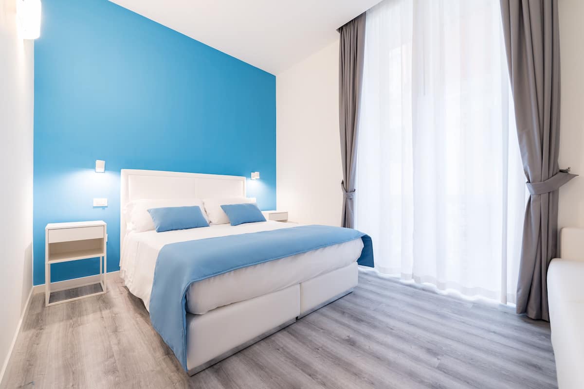 Une chambre à coucher moderne, dotée d’un grand lit, de tables de chevet et offrant une vue sur l’extérieur à travers des rideaux légers.