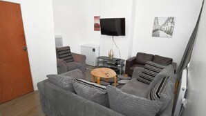Moderni olohuone, jossa on sohva, nojatuoleja, sohvapöytä, taulutelevisio ja pieni jääkaappi.