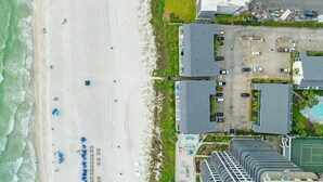 Vue aérienne d’un hôtel situé sur la côte, avec une plage, un parking et plusieurs bâtiments.