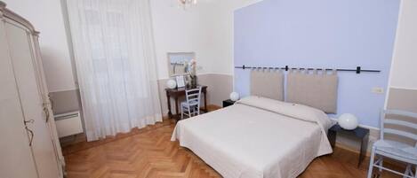 A bedroom with a wooden herringbone floor, a bed with a beige bedspread, a nightstand with a lamp, a chair, and a wardrobe.