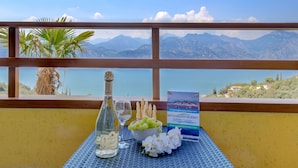 A table with a bottle of wine, glasses, a bowl of grapes, and a brochure, set against a scenic mountain view. - Garden Residence Malcesine Lake View (Malcesine)