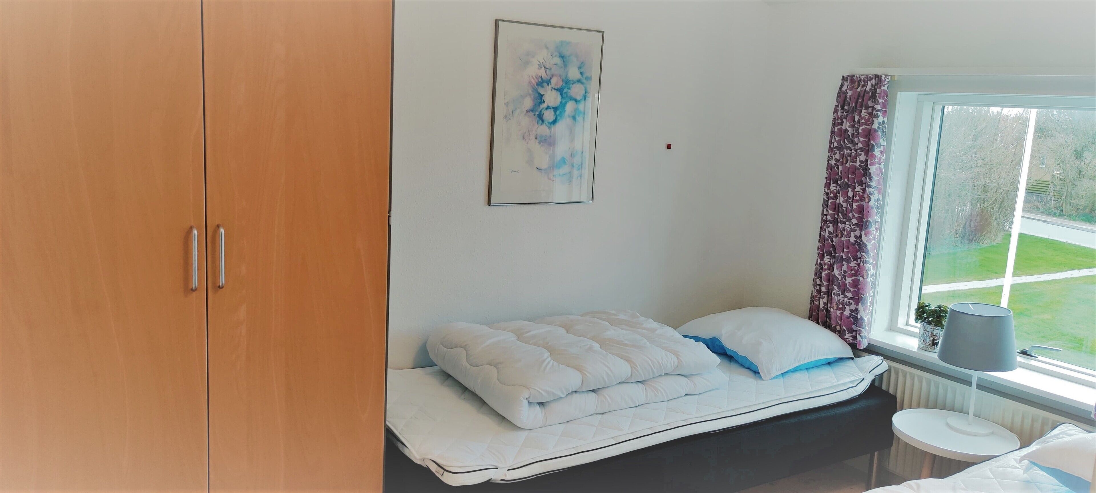 Een slaapkamer met een bed, een kledingkast, een raam met gordijnen en een schilderij aan de muur.