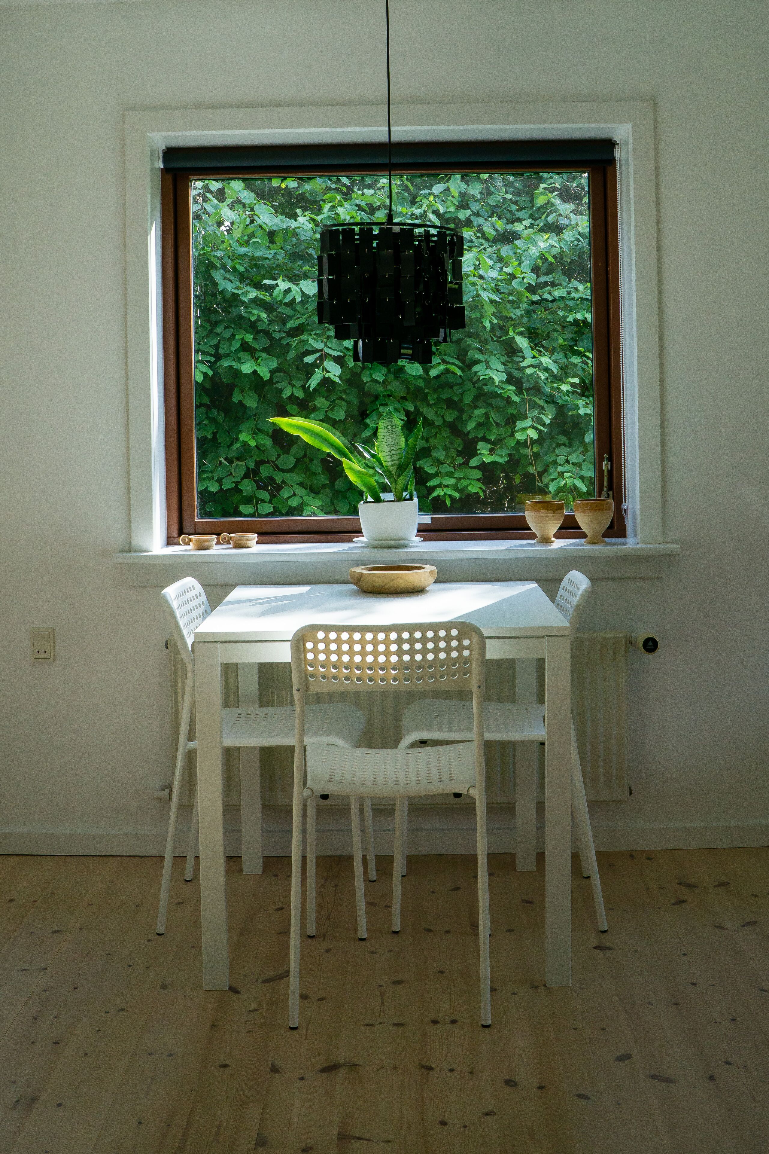 A dining area with a white table and chairs, a window with a view of greenery, and a hanging light fixture.