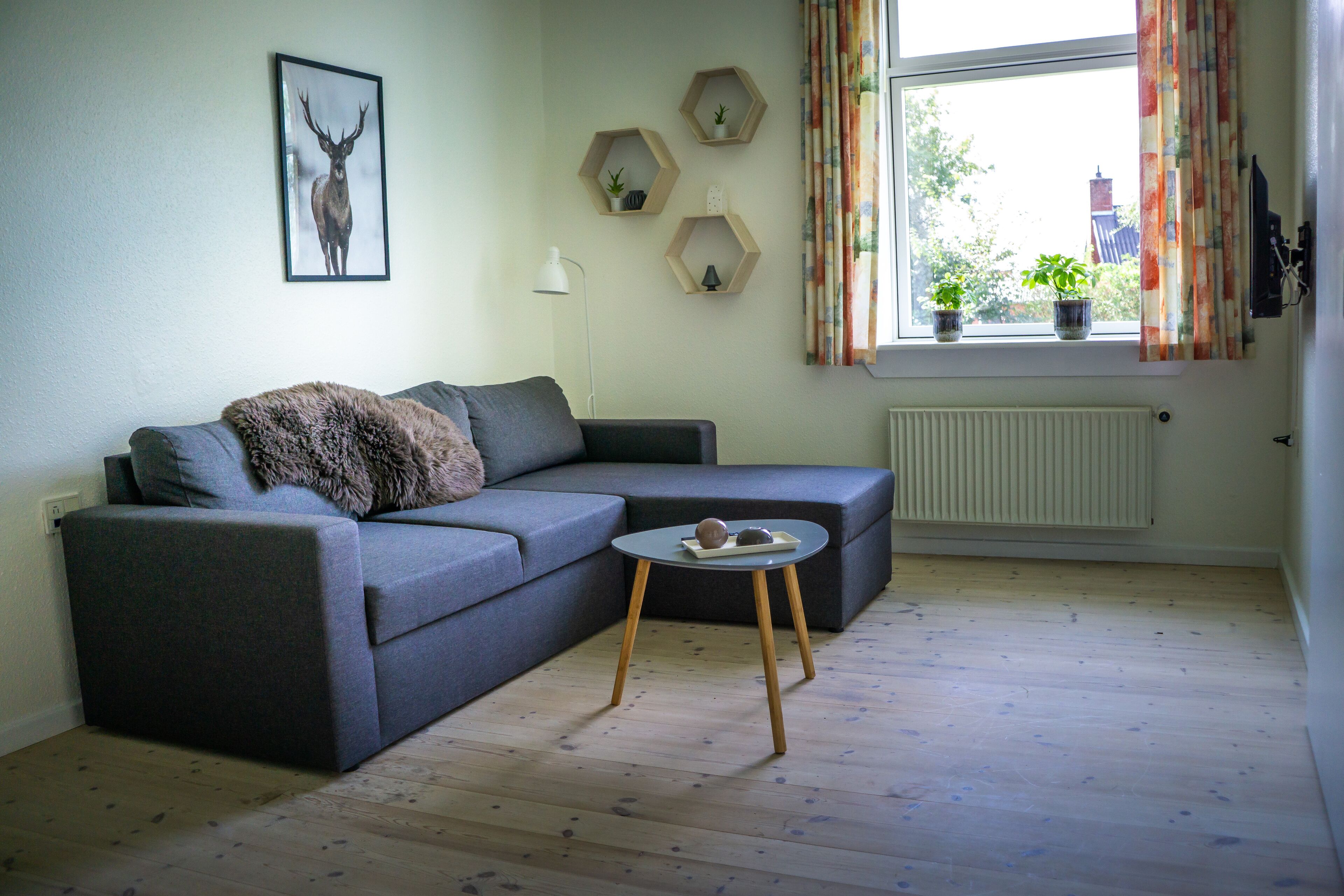 Un salon moderne avec un canapé d’angle gris, une petite table ronde agrémentée de deux objets décoratifs, un tableau représentant un cerf encadré au mur, et une fenêtre avec des rideaux et des plantes en pot sur le rebord.