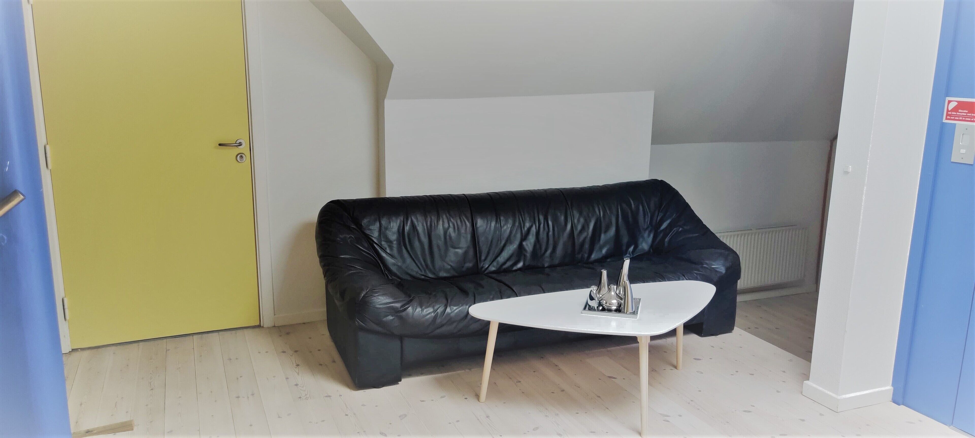 Een kamer met een zwarte leren bank, een witte salontafel, een gele deur en een witte radiator.