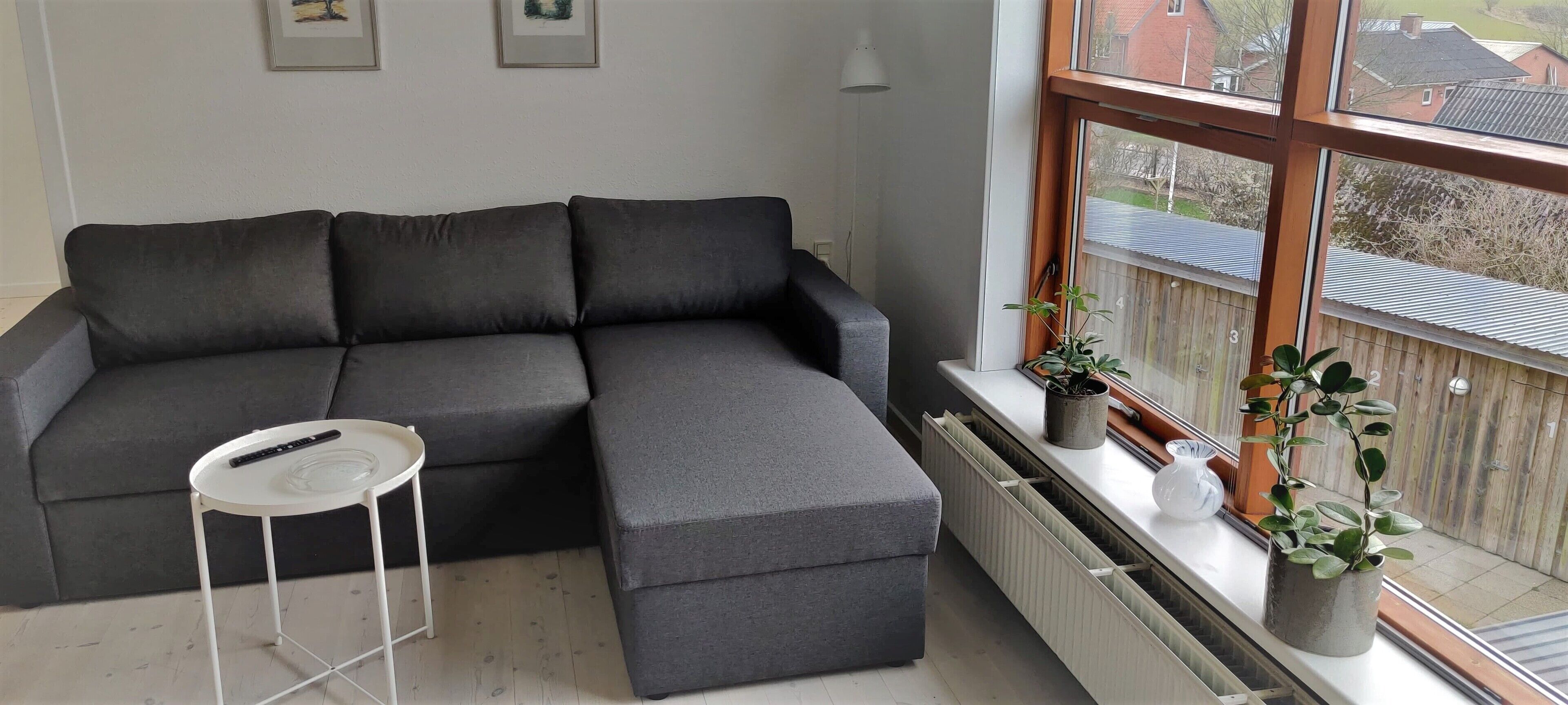 En grå hjørnesofa med et lille hvidt sengebord, en radiator og potteplanter ved et vindue.