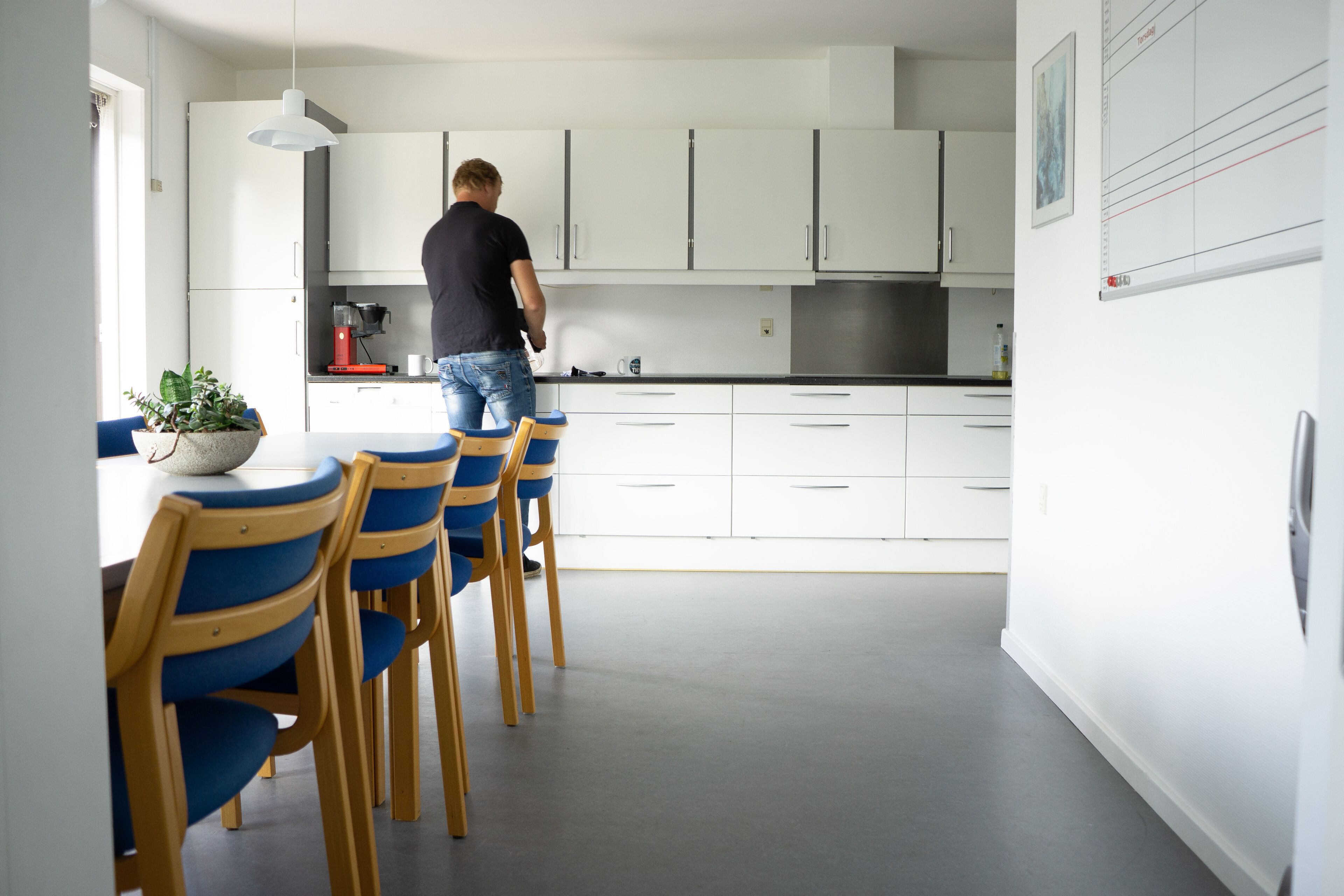 Une cuisine moderne avec des meubles blancs, un coin repas avec des chaises bleues, et une personne debout près du comptoir.