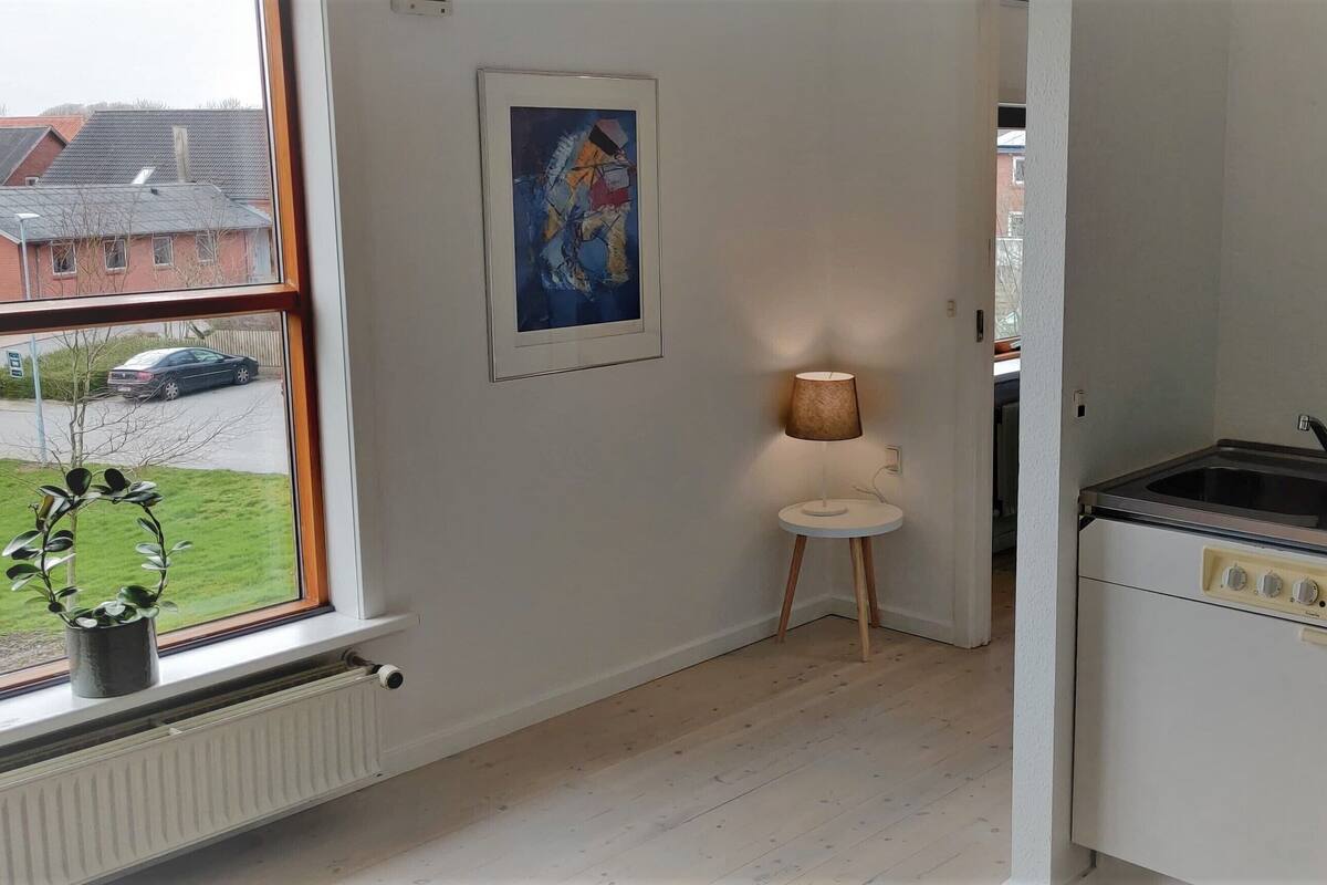 A compact kitchen with a built-in dishwasher, a small table with a lamp, and a window with a view of a street and houses.