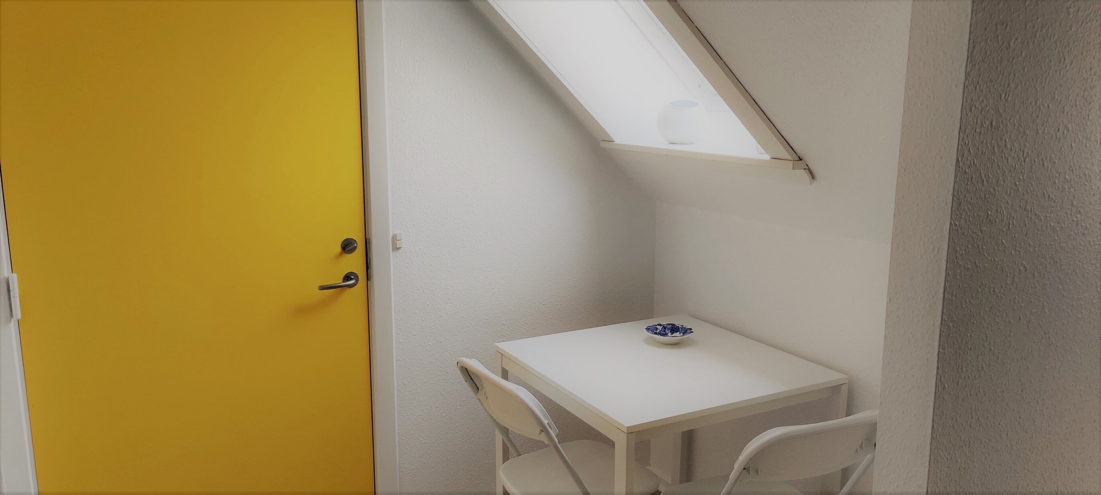 A small room with a yellow door, a white table with a blue bowl, and a white chair.
