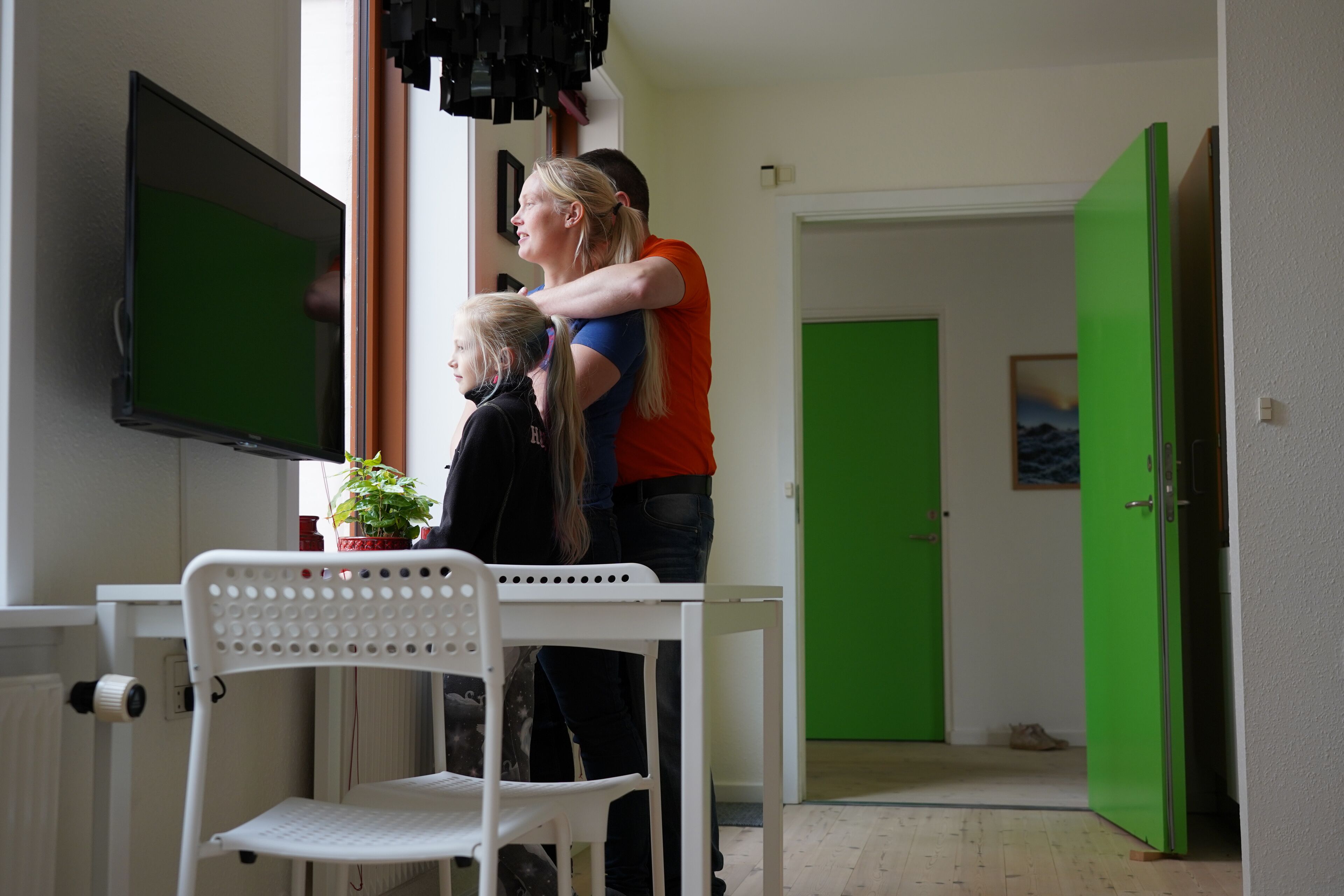 Une pièce avec une chaise blanche, une table sur laquelle est posée une plante en pot, une porte verte et un téléviseur fixé au mur.