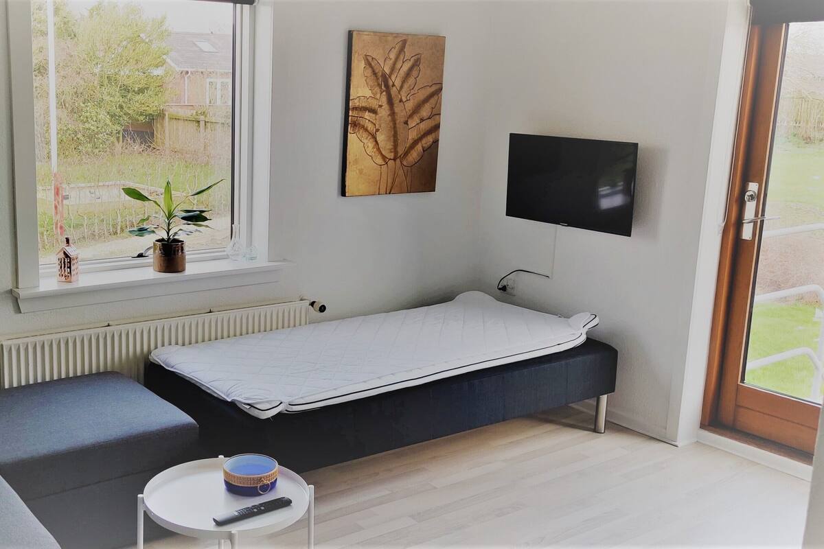 A compact room with a single bed, a small table, a chair, a desk lamp, a TV, and a window with a view of the outdoors.
