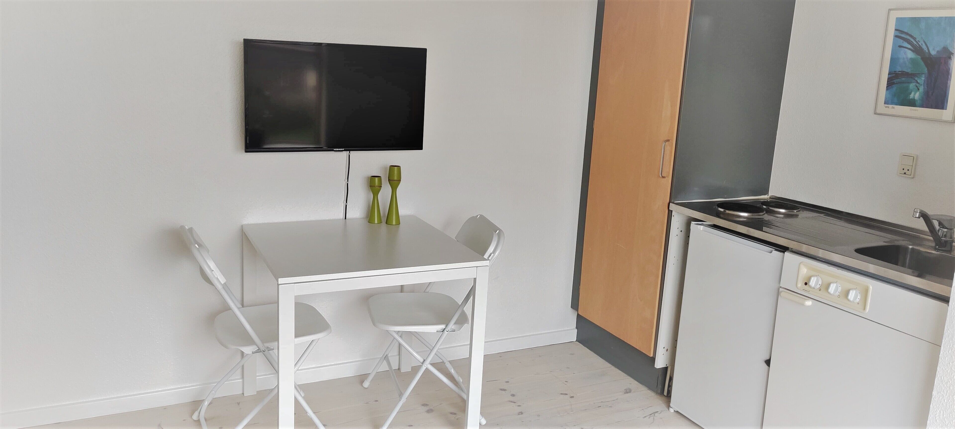 A compact kitchen with a white table and chairs, a flat-screen TV mounted above, and a small cabinet with a sink.