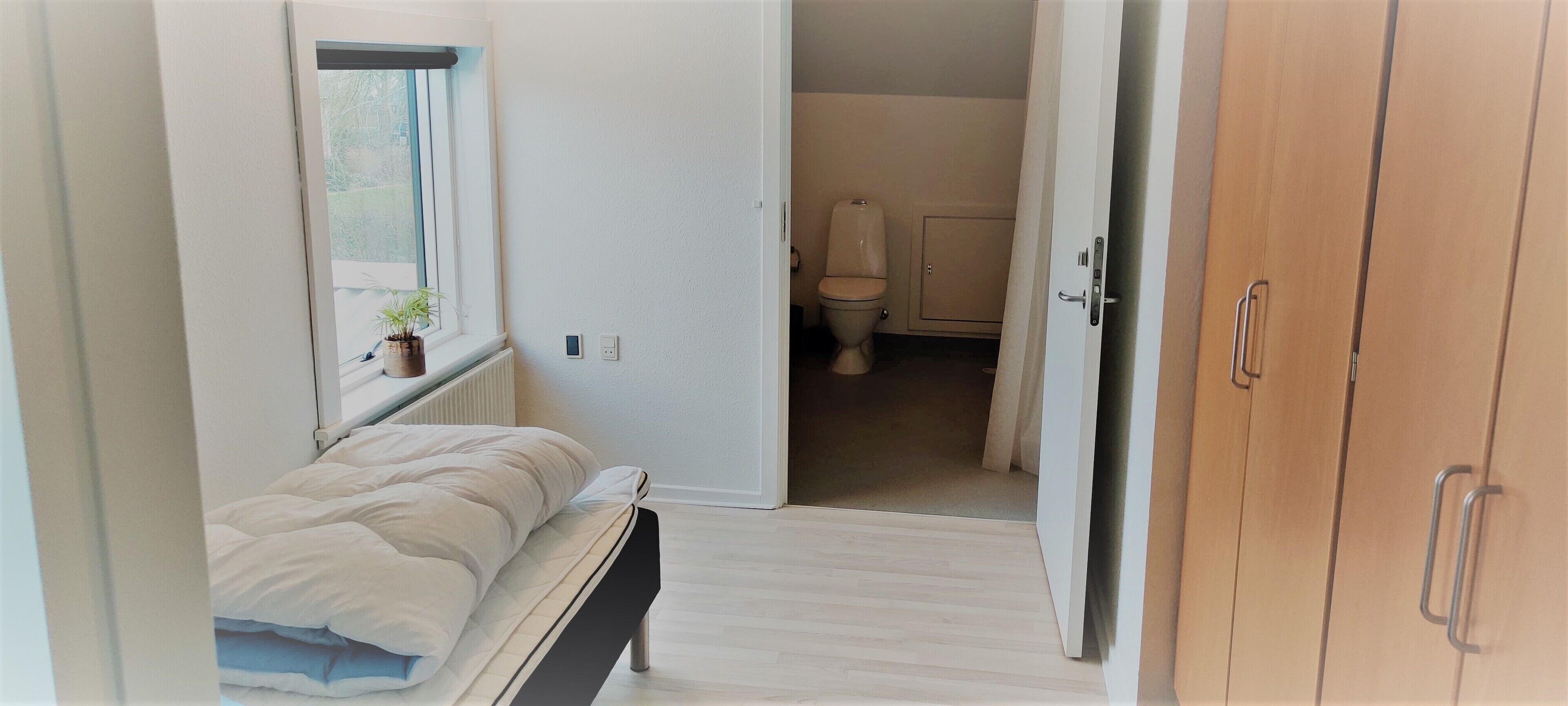 Ein Schlafzimmer mit einem Bett, einem Fenster mit Pflanze, einer weißen Tür, die ins Badezimmer mit Toilette und Schrank führt.