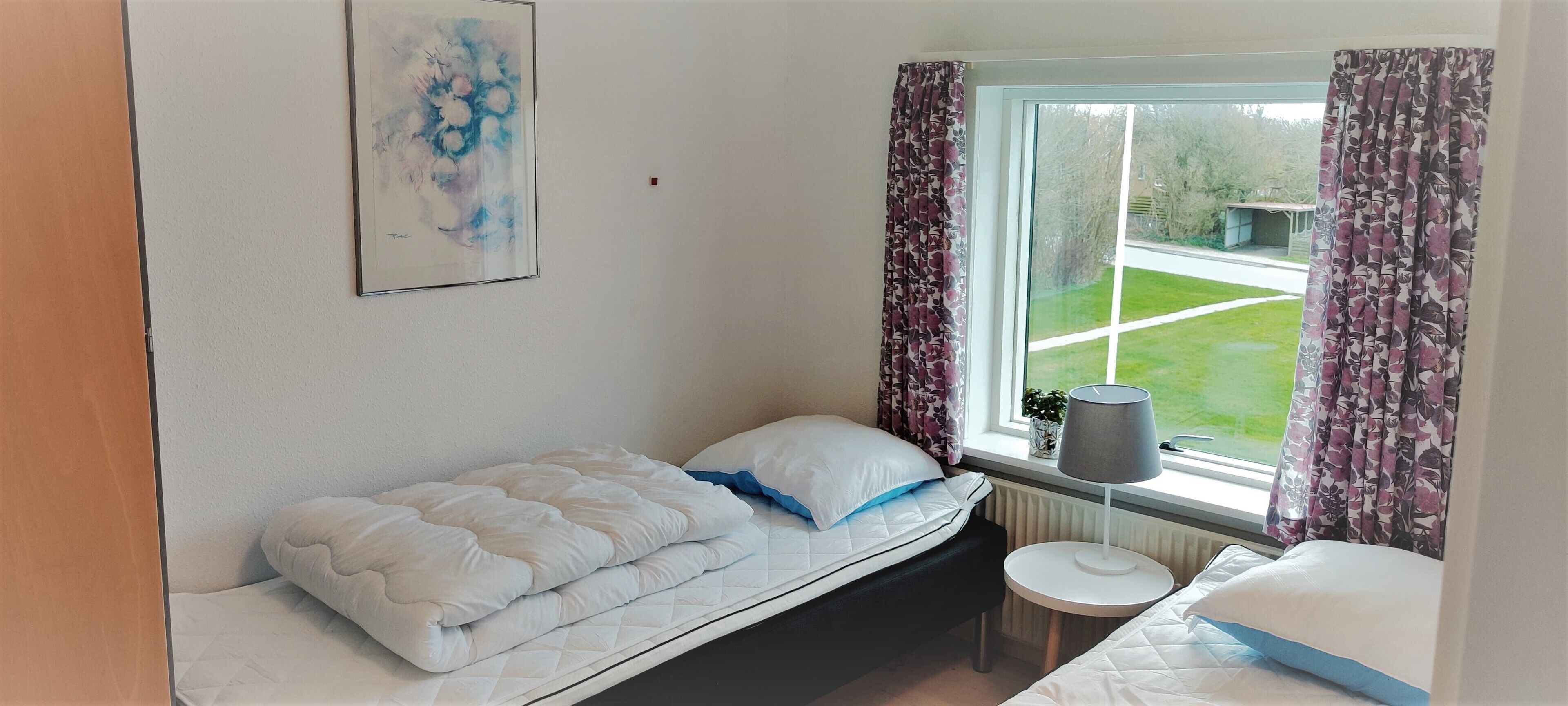 A bedroom with a bed, a small round table, a window with curtains, and a painting on the wall.