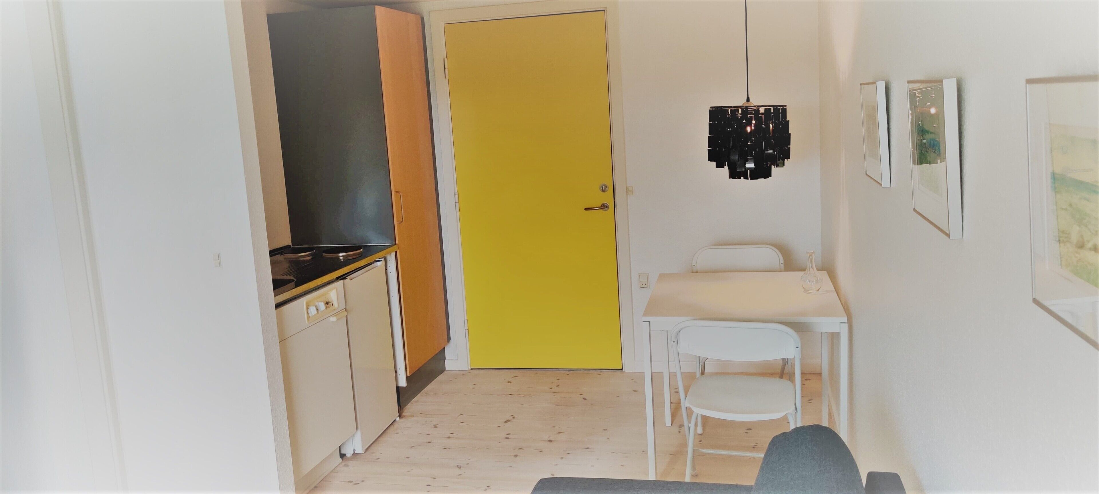 A compact kitchen with a yellow door, a white dining area with a small table and chairs, and a hanging light fixture.
