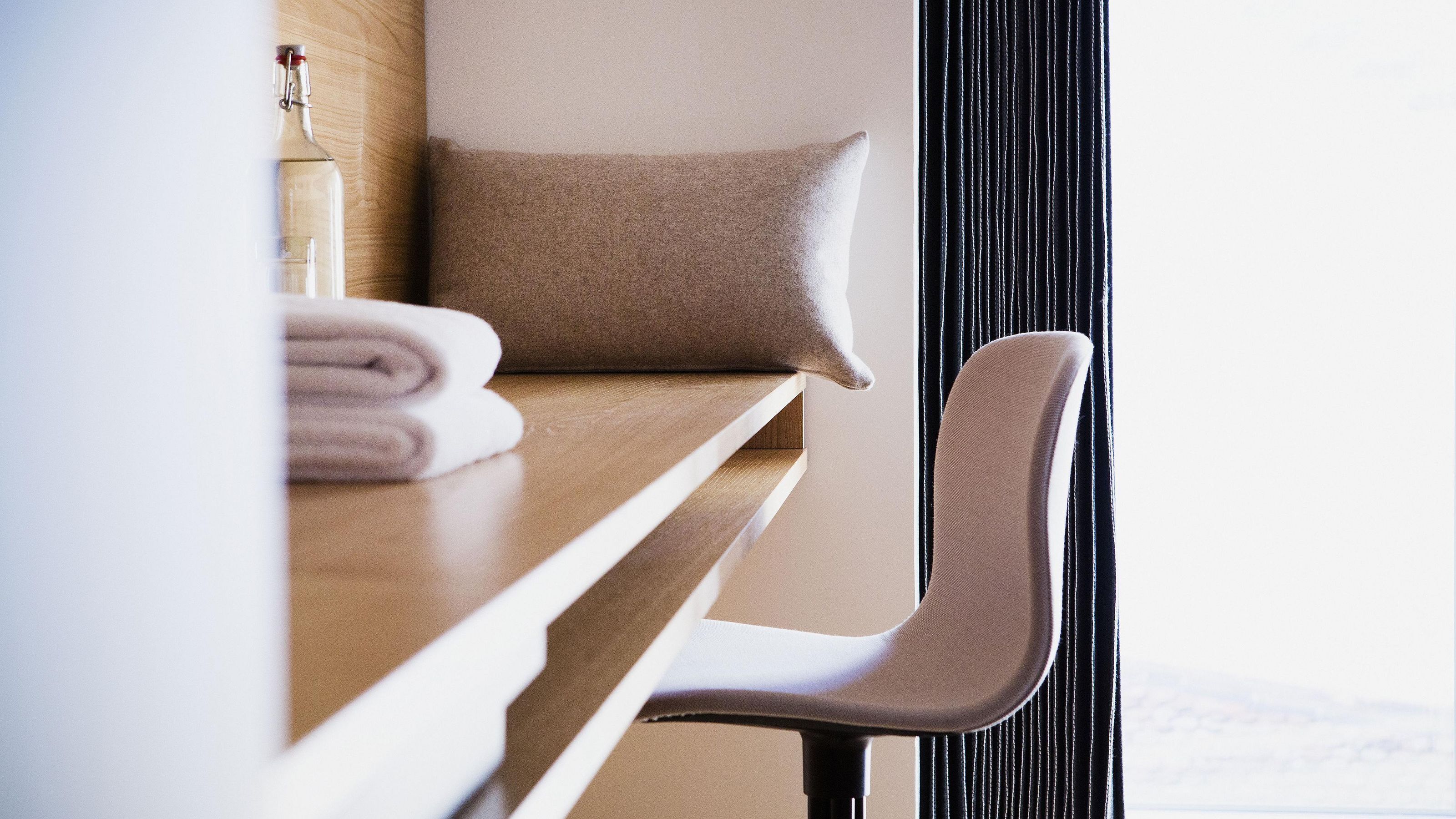 A neatly made bed with a folded towel and a pillow, a wooden bedside table, and a chair with a headrest.