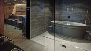 A modern bathroom with a freestanding bathtub, dark tiled walls, and a wooden bench area. - Boutique Hotel Schlossberg (Thun)