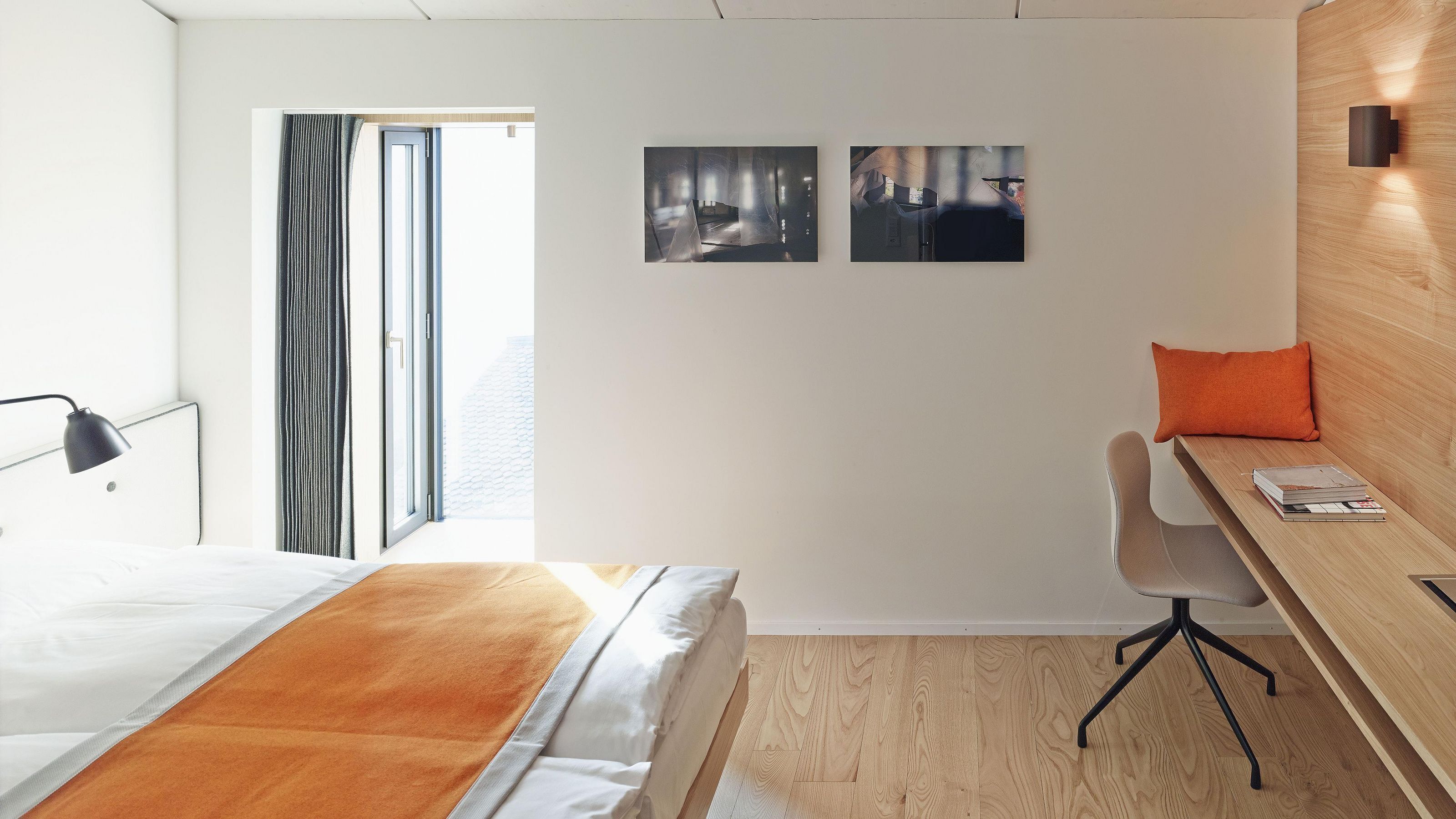 A modern hotel room with a bed, a desk, and a chair. There are two wall-mounted pictures and a window with curtains.
