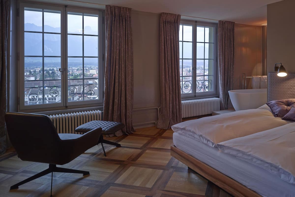 Una habitación de hotel con una cama grande, un escritorio y vistas a un paisaje urbano a través de amplias ventanas.
