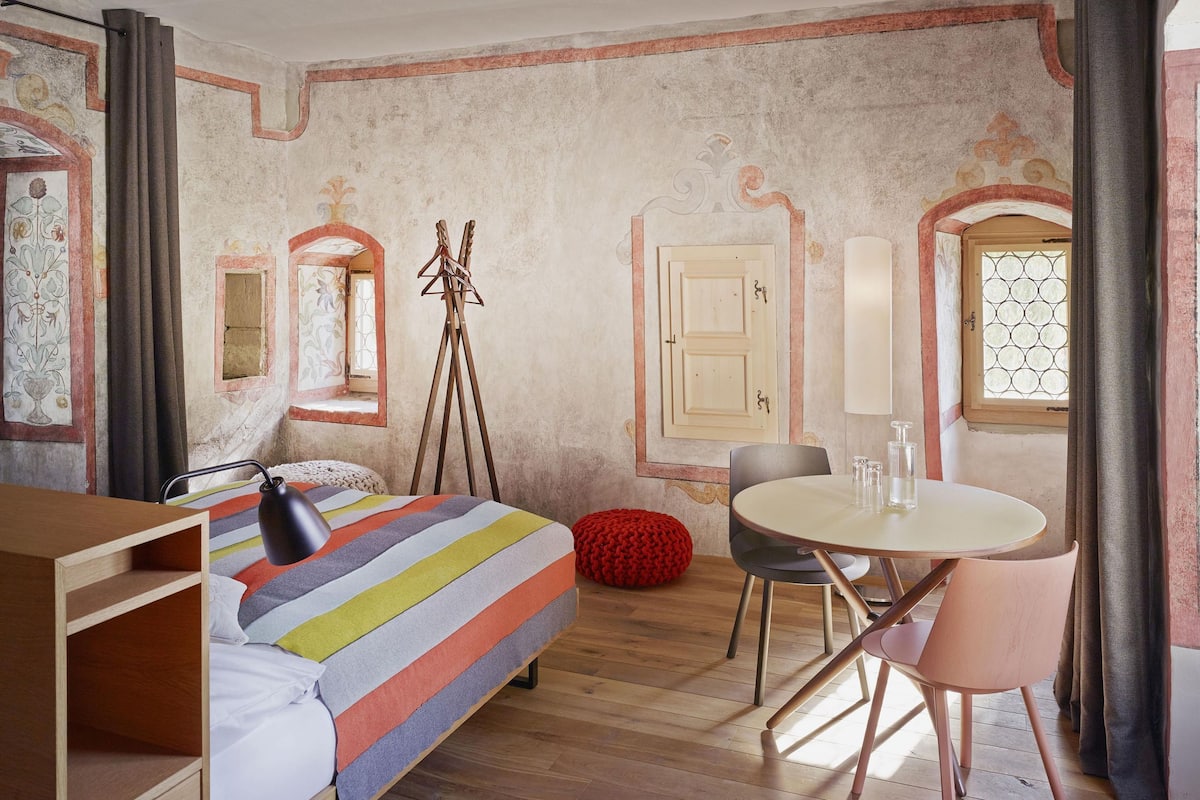 Una habitación con una cama, una mesita pequeña con sillas, un taburete rojo y un espejo.