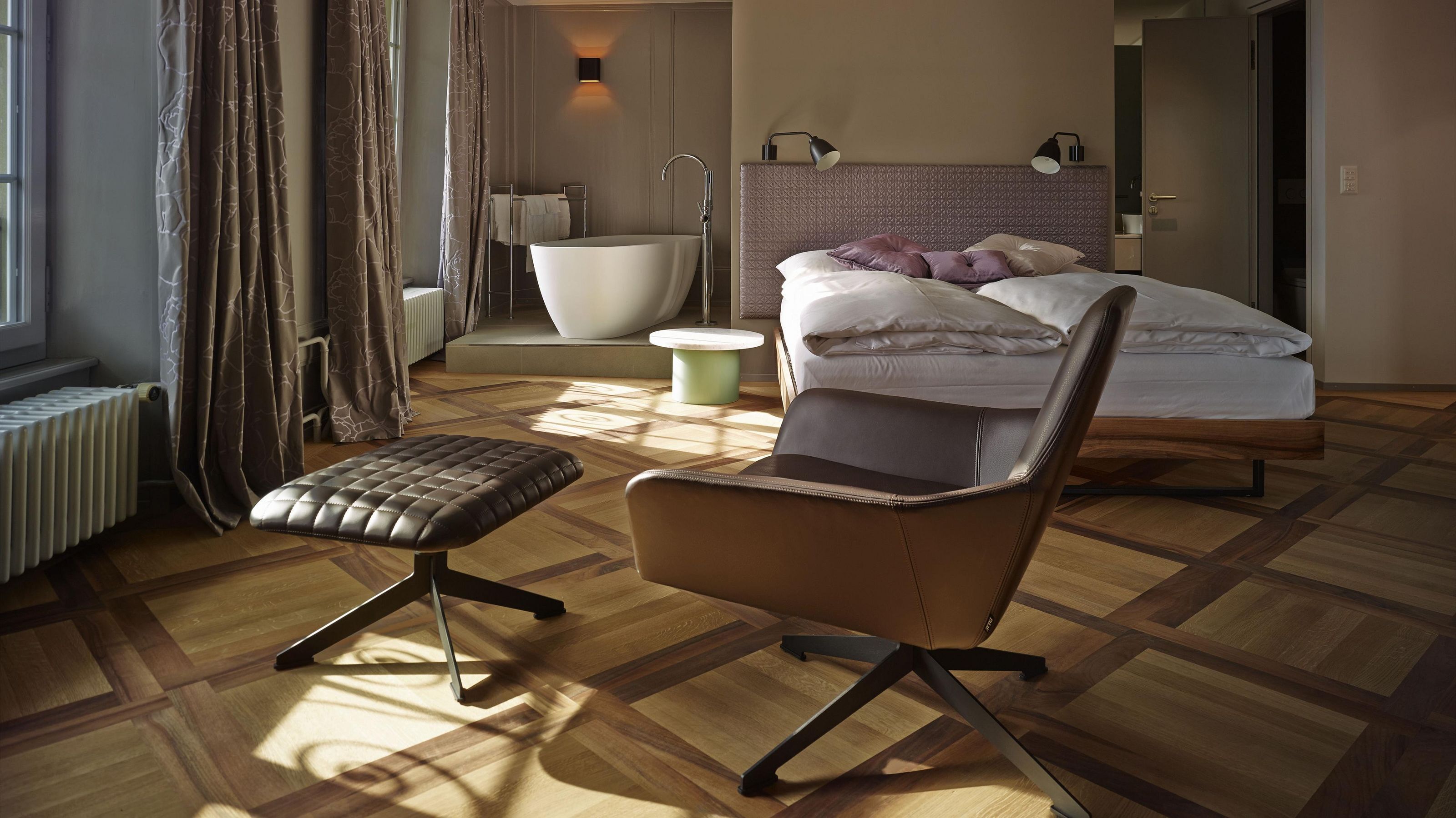 A modern hotel room with a wooden parquet floor, a large bed with a headboard, a sitting area with a cushioned chair, a small round table, and a bathtub.
