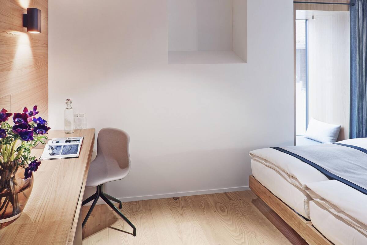Una habitación de hotel moderna con suelo de madera, un escritorio con silla, una cama con cabecera azul y una ventana con cortinas.