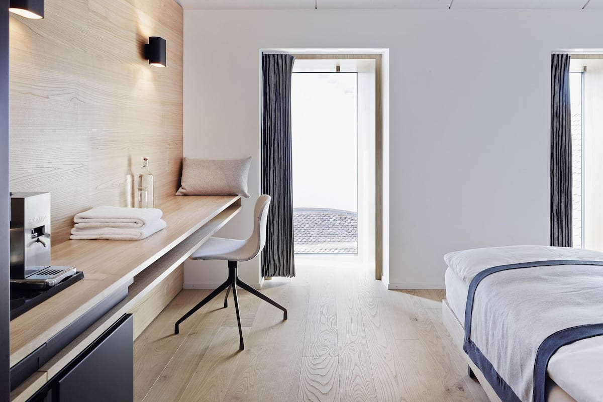 Habitación de hotel con escritorio de madera, una silla, una cama y vista a un muro de ladrillo a través de una ventana.