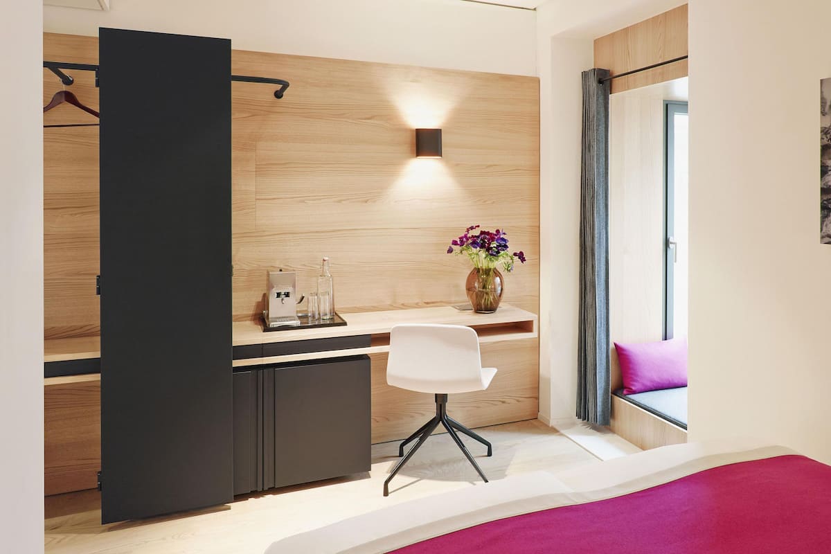 Un dormitorio moderno con cama, escritorio, silla, un jarrón con flores y ventana con vistas.