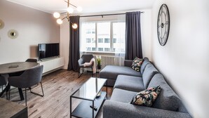 Una sala moderna con un sofá gris, una mesa de centro de vidrio, una mesa de comedor con sillas, un televisor sobre un mueble bajo y un gran ventanal con cortinas.