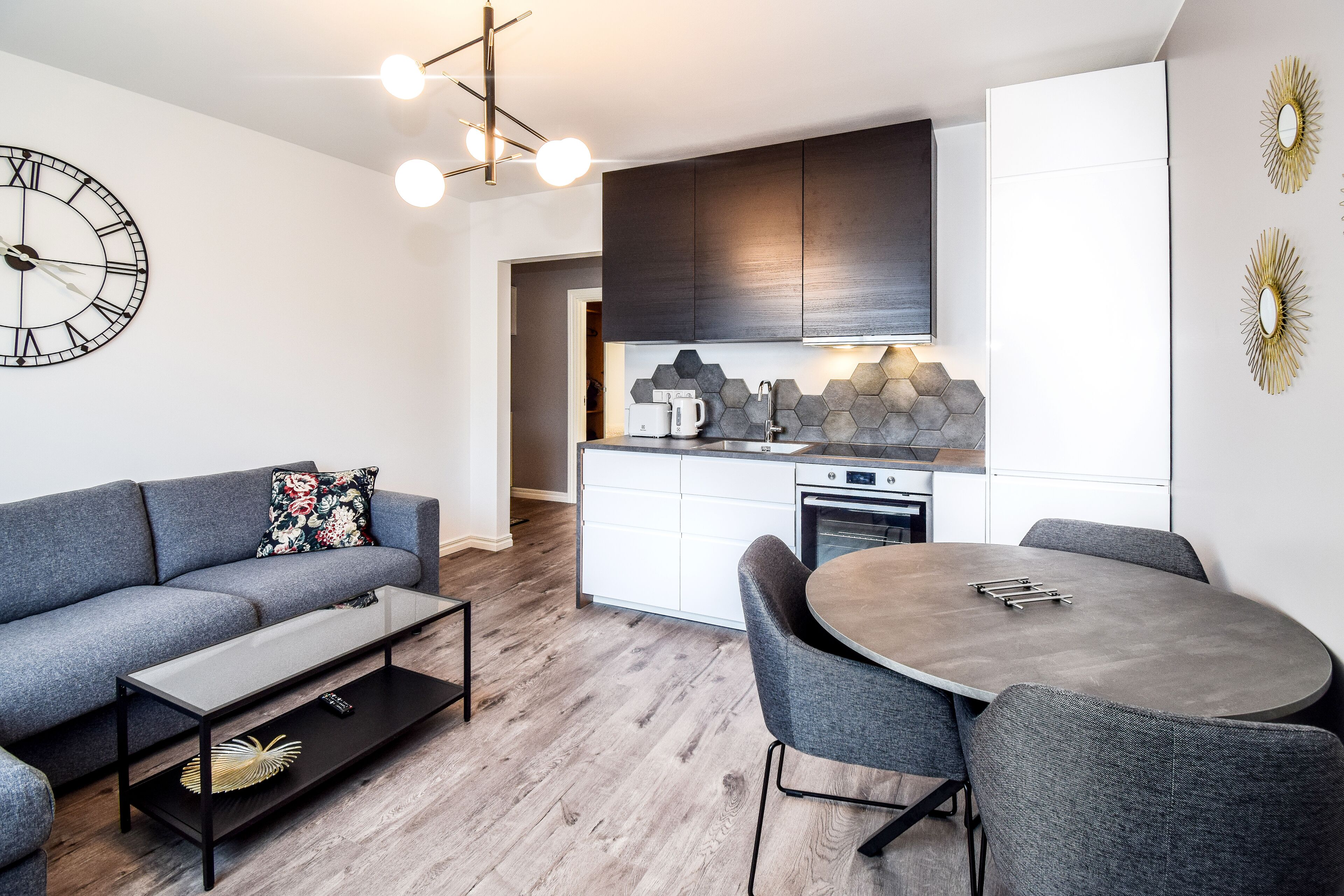 Een moderne woonkamer met een grijze bank, een ronde eettafel met grijze stoelen, een koffieapparaat en een keuken met donkere kastjes en een zeshoekige achterwand.