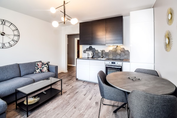 A modern living room with a grey sofa, a round dining table with grey chairs, a coffee machine, and a kitchen with dark cabinets and a hexagonal backsplash.