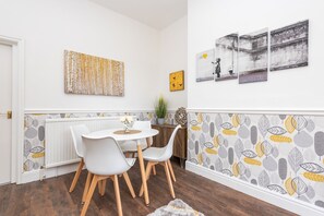 Un coin repas avec une table et des chaises blanches, un radiateur en dessous et un papier peint à motifs de feuilles.