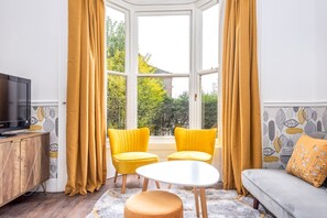 Une pièce avec une grande fenêtre en saillie, deux fauteuils jaunes, une table blanche, un canapé et un téléviseur posé sur un meuble en bois.