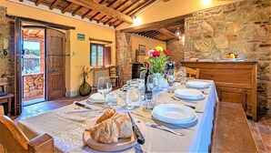 Une table à manger dressée avec des assiettes, des verres et un pain, dans une pièce aux poutres apparentes et aux murs en pierre.