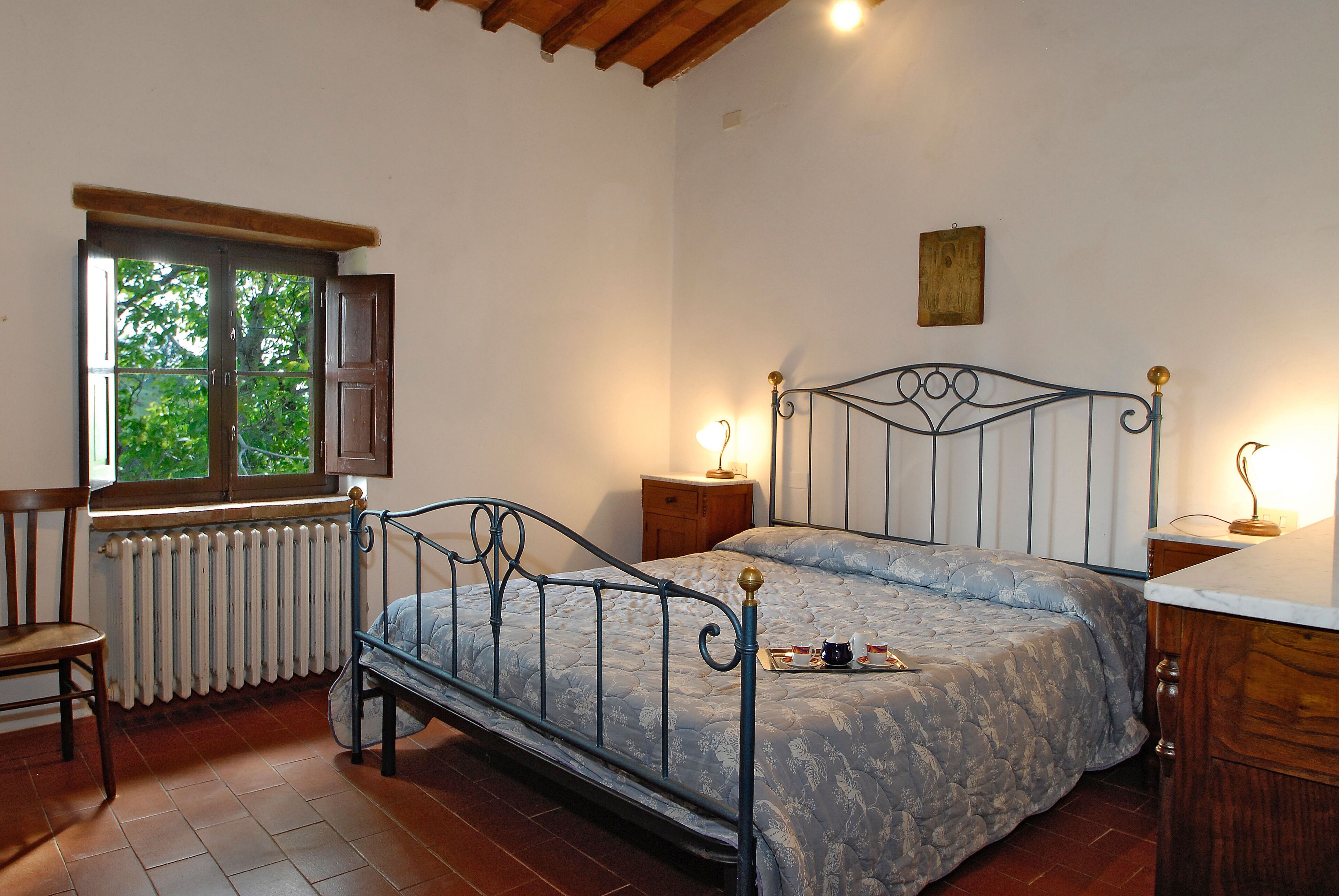 A bedroom with a metal bed frame, a wooden chair, a radiator, a window with a view of greenery, and bedside lamps.